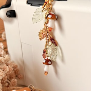 May include: A gold-toned metal charm featuring two small mushrooms with white stems and brown caps with white spots, interspersed with translucent pale green leaf-shaped beads, small amber-colored crystals, and textured metallic leaf accents.