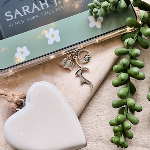 Op de afbeelding: Een wit, hartvormig ornament van keramiek met een touwtje eraan. Het ornament ligt op een beige stoffen oppervlak met een boekomslag op de achtergrond. De boekomslag is groen en heeft de tekst "SARAH J. MAAS #1 NEW YORK TIMES BESTSELLING AUTHOR" en een bloemenpatroon.