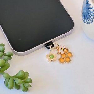 May include: A gold charm with a bee, a flower, and a honeycomb design. The charm is attached to a black phone case.