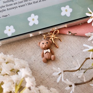Könnte beinhalten: Ein brauner Teddybär-Anhänger mit weißer Fliege und goldenem Schleifenakzent. Der Anhänger ist an einer transparenten Handyhülle befestigt. Der Hintergrund umfasst ein Buch und weiße Blumen.