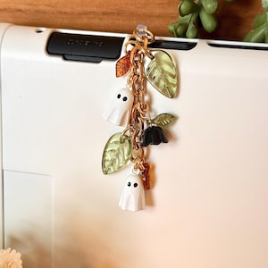 May include: A gold-toned metal chain charm featuring two white ghost pendants with black eyes, translucent green leaf charms, a small black flower charm, and a small amber-colored leaf charm, all attached to a circular jump ring connector.