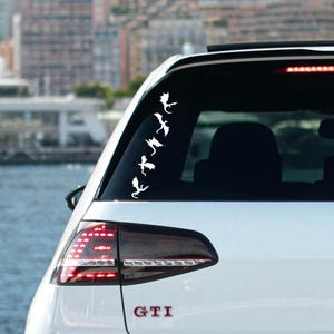 May include: White car with a black rear window and a white decal of a line of dragons on the back window. The car has a red taillight and the word "GTI" on the back bumper.