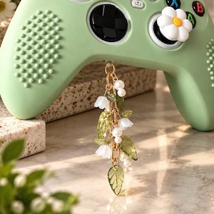 May include: A light green video game controller featuring a white daisy-shaped joystick cover and a dangling charm. The charm consists of a gold-toned chain adorned with small white pearls, translucent green leaf-shaped beads, and white bell-shaped flower beads.