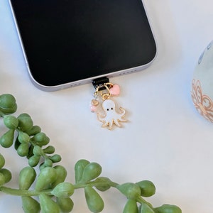 May include: A white octopus charm with pink hearts hanging from a black phone charger. The octopus has black eyes and a gold outline.