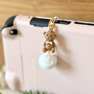 May include: A brown teddy bear charm with a white bow tie and a fluffy white pom-pom, attached to a gold-colored chain. The charm is hanging from a pink electronic device.