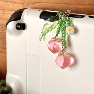 May include: A white handheld gaming device with a peach-themed charm. The charm features two pink, translucent peach pendants, green beads, a white daisy, and a green leaf. The charm is attached to the device with a silver chain.