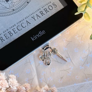 May include: A black Kindle e-reader with a clear case and a silver dragon and sword charm attached. The Kindle is on a light pink fabric with a floral pattern.