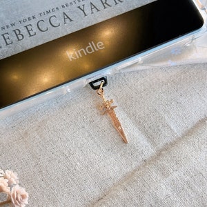Puede incluir: Un marcapáginas de espada dorado con un mango decorativo, colgando de un lector electrónico Kindle con una funda transparente. La pantalla del lector electrónico muestra el texto "NEW YORK TIMES BESTSELLING AUTHOR REBECCA YARROS" y la palabra "kindle".