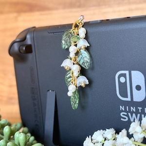 Peut inclure: Un charm doré orné de fleurs de muguet, de feuilles vertes et de perles blanches. Le charm est fixé à une console Nintendo Switch noire. Le design comprend de petites fleurs blanches en forme de cloche et des feuilles vertes détaillées.