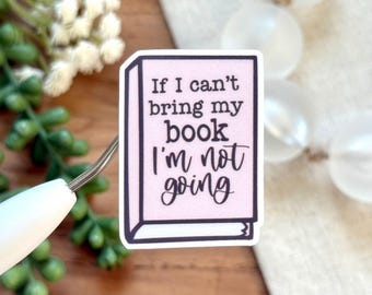 May include: A rectangular sticker with a light pink background and black border. The sticker reads "If I can't bring my book I'm not going" in a mix of fonts. The sticker is held by a tool against a wooden surface.
