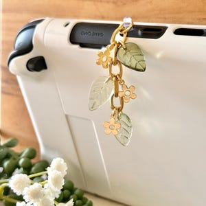 May include: A gold-toned charm with clear leaf and flower pendants is attached to a white handheld gaming device. The charm is suspended from a small hook. The words "GAME CARD" are visible on the device.