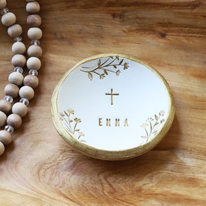 May include: White ceramic ring dish with a gold rim and floral design. The dish has a gold cross and the name "EMMA" written in gold lettering.