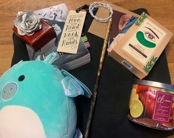 May include: A selection of items including a teal dragon plush, a red jewelry box, a bookmark with text, a wand, a candle, and eye treatments. The candle has a tropical sangria label. The bookmark reads "...may you always have love in your heart and a book in your hands."