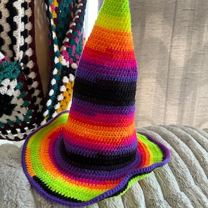May include: A colorful crocheted witch hat with a wide brim and a pointed crown. The hat is striped with black, green, orange, pink, and purple yarn.