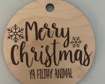 Op de afbeelding: Een ronde houten ornament met de tekst "Merry Christmas Ya Filthy Animal" en een sneeuwvlokontwerp.