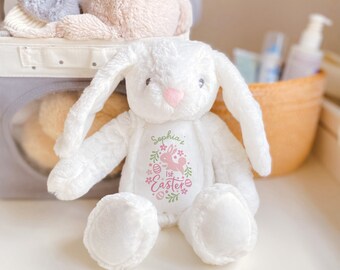 May include: A white plush bunny rabbit with long floppy ears and a pink nose. The bunny has a personalized design on its chest that reads "Sophia's 1st Easter" with a pink bunny and floral accents. Other plush toys are in the background.