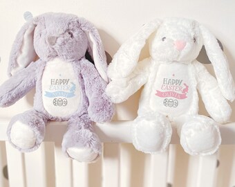 May include: Two plush Easter bunnies, one lavender and one white, sit side-by-side. Each bunny has long floppy ears and a stitched face. The lavender bunny has "Happy Easter" and the name "Noah" embroidered on its chest. The white bunny has "Happy Easter" and the name "Olivia".