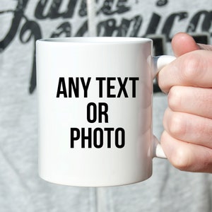 Può includere: Una tazza di ceramica bianca con manico, tenuta da una persona. La tazza presenta le parole "ANY TEXT OR PHOTO" stampate in grassetto nero. Lo sfondo è grigio chiaro.