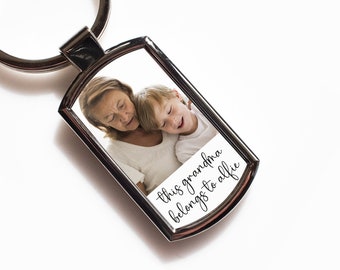 May include: Silver rectangular keychain with a photo of a woman and a child. The photo is framed by a silver metal border. The text "this grandma belongs to alfie" is printed on the photo.