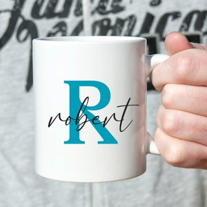 Può includere: Tazza in ceramica bianca con la lettera "R" turchese e il nome "robert" in corsivo nero. La tazza è tenuta da una persona. Lo sfondo è grigio chiaro.