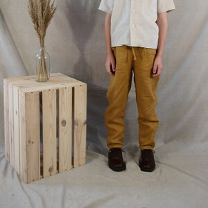 Puede incluir: Una persona joven lleva una camisa de lino beige claro con mangas cortas y cuello abotonado. Tambi&eacute;n lleva pantalones de lino amarillo mostaza y zapatos de cuero marr&oacute;n. La persona est&aacute; de pie frente a un fondo beige claro con una caja de madera y un jarr&oacute;n de hierbas secas.