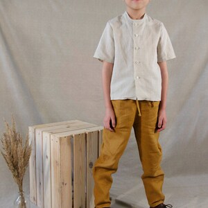Puede incluir: Una persona joven lleva una camisa de lino beige claro con cuello mandar&iacute;n y mangas cortas, combinada con pantalones de lino amarillo mostaza y zapatos de cuero marr&oacute;n. La persona est&aacute; de pie frente a una caja de madera.
