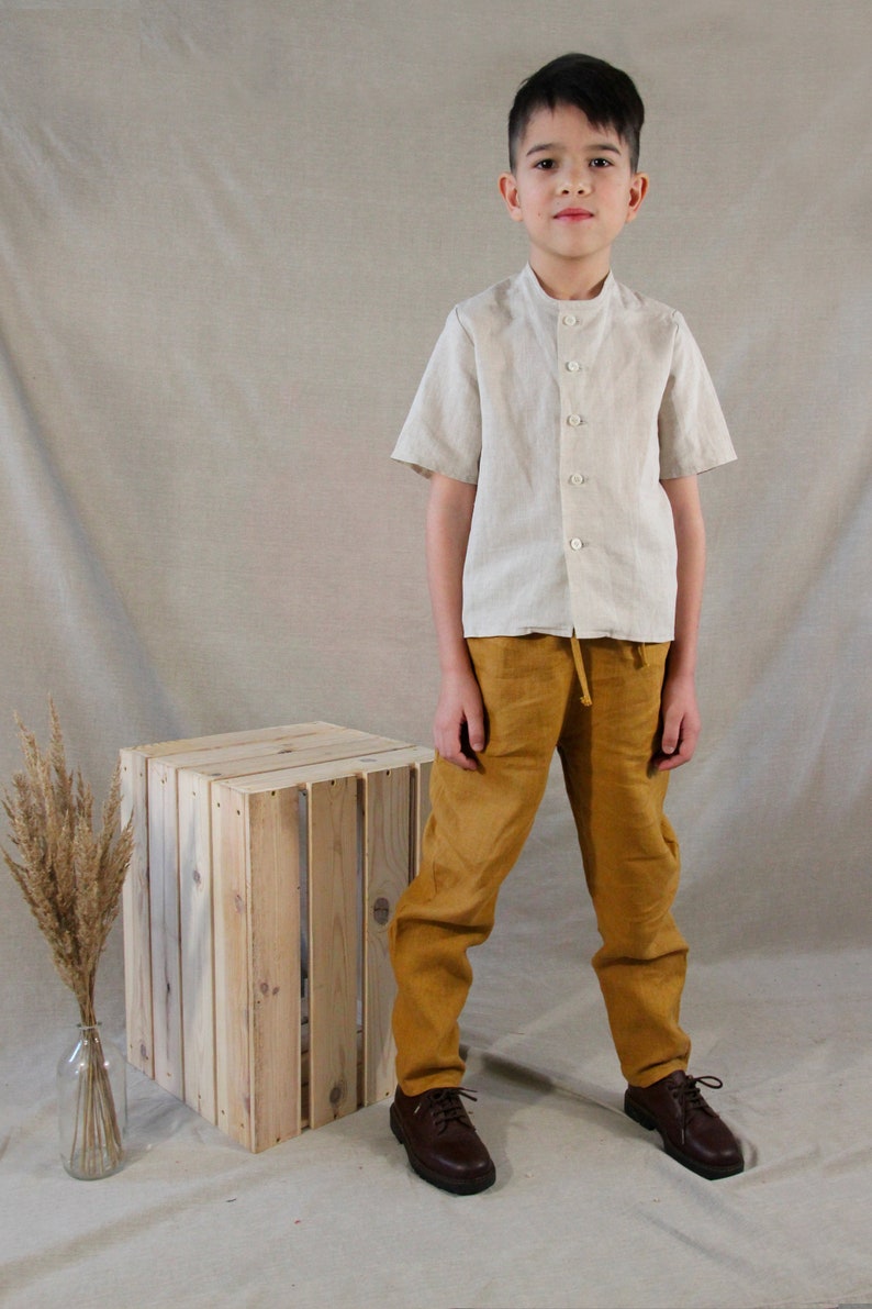 Puede incluir: Una persona joven lleva una camisa de lino beige claro con cuello mandar&iacute;n y mangas cortas, combinada con pantalones de lino amarillo mostaza y zapatos de cuero marr&oacute;n. La persona est&aacute; de pie frente a una caja de madera.