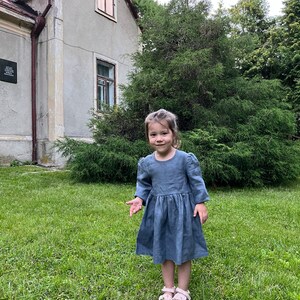 May include: A young girl wearing a light blue linen dress with puffed sleeves stands in a grassy area in front of a house. She is smiling and looking at the camera.