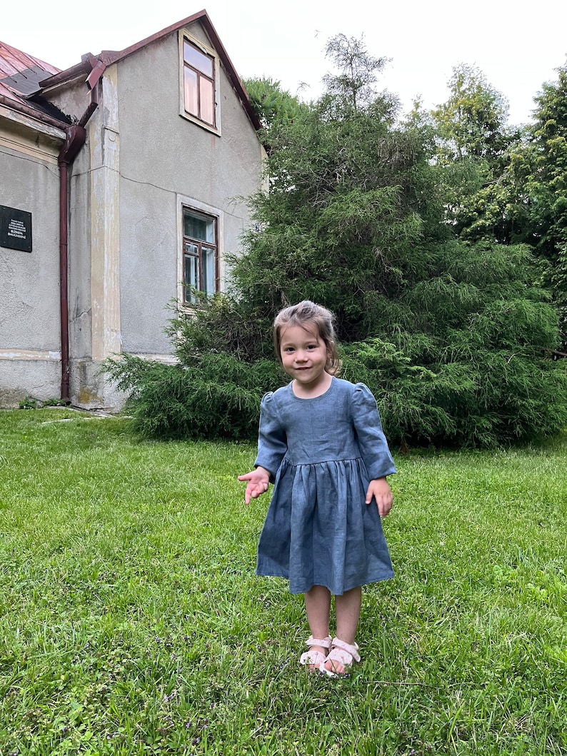 May include: A young girl wearing a light blue linen dress with puffed sleeves stands in a grassy area in front of a house. She is smiling and looking at the camera.