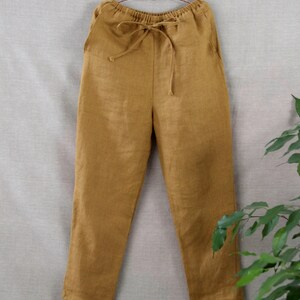 Puede incluir: Pantal&oacute;n de lino amarillo mostaza con cintura de cord&oacute;n. El pantal&oacute;n est&aacute; colgado de una percha marr&oacute;n sobre un fondo gris claro.