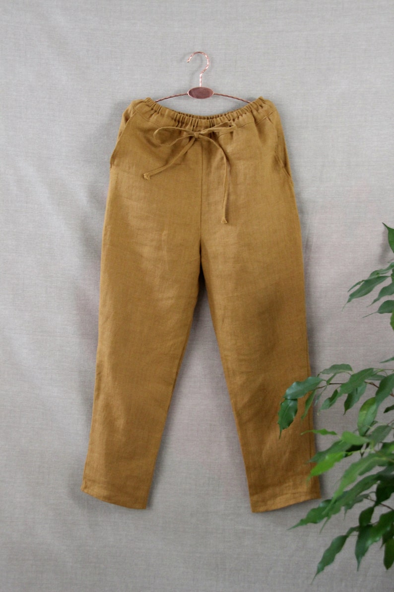 Puede incluir: Pantal&oacute;n de lino amarillo mostaza con cintura de cord&oacute;n. El pantal&oacute;n est&aacute; colgado de una percha marr&oacute;n sobre un fondo gris claro.