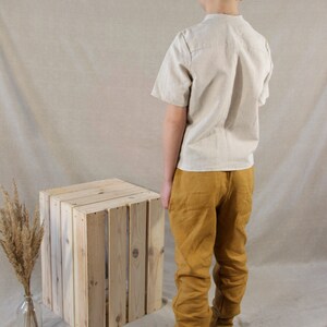 Puede incluir: Una persona joven lleva una camisa de lino beige claro y pantalones de lino amarillo mostaza. Est&aacute; de pie frente a una caja de madera y lleva zapatos de cuero marr&oacute;n.