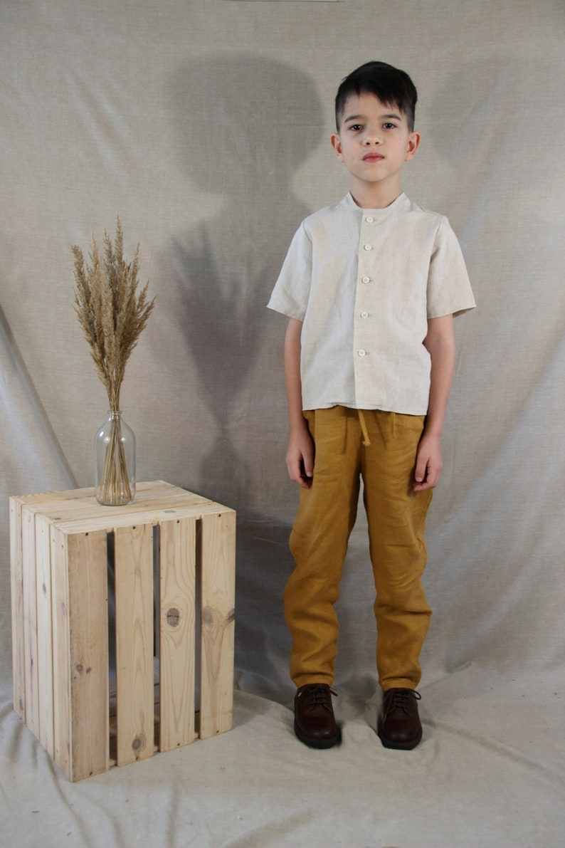 Puede incluir: Una persona joven lleva una camisa de lino beige claro con mangas cortas y cuello abotonado. Tambi&eacute;n lleva pantalones de lino amarillo mostaza y zapatos de cuero marr&oacute;n. La persona est&aacute; de pie frente a un fondo beige claro con una caja de madera y un jarr&oacute;n de hierbas secas.