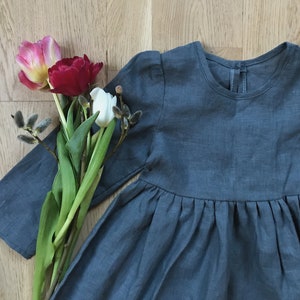 May include: A gray linen dress with long sleeves and a gathered skirt. The dress is laying on a wooden surface with a bouquet of tulips and pussy willows.