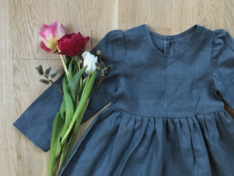 May include: A gray linen dress with long sleeves and a gathered skirt. The dress is laying on a wooden surface with a bouquet of tulips and pussy willows.