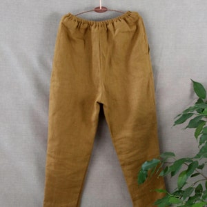 Puede incluir: Un pantal&oacute;n de lino marr&oacute;n con cintura de cord&oacute;n. El pantal&oacute;n est&aacute; colgado en una percha de madera sobre un fondo gris claro.