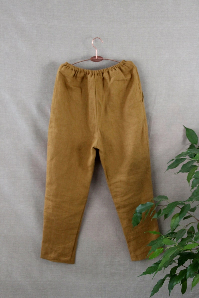 Puede incluir: Un pantal&oacute;n de lino marr&oacute;n con cintura de cord&oacute;n. El pantal&oacute;n est&aacute; colgado en una percha de madera sobre un fondo gris claro.