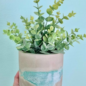 May include: A concrete planter with a light blue and beige color scheme, holding a faux eucalyptus plant. The cylindrical pot has a textured surface and a band of turquoise paint. The plant has green leaves and stems.