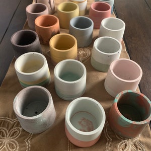 May include: A collection of small, cylindrical concrete planters in various colors, including pink, yellow, and gray. Some planters have painted stripes or color-blocked designs. The planters are arranged on a brown surface.