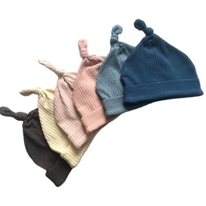 May include: A collection of ribbed knit baby hats in various colors, including dark brown, cream, pink, light blue, and navy blue. Each hat features a knotted top and a folded brim, perfect for newborns.