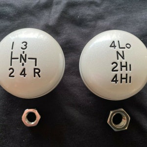 May include: Two silver gear shift knobs with black lettering. The left knob has the numbers 1, 2, 3, 4, and R. The right knob has the letters 4Lo, N, 2Hi, and 4Hi.