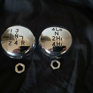 May include: Two chrome gear shift knobs with black lettering. The left knob has the numbers 1, 2, 3, 4, and R, and the letters H and N. The right knob has the numbers 4, 2, and the letters L, O, N, H, and I.