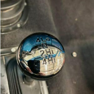 May include: A chrome gear shifter with a blue reflective surface and the following markings: 4L, N, 2H, 4H.