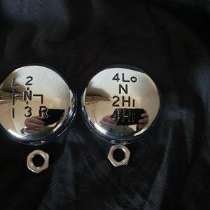 May include: Two chrome gear shift knobs with black lettering. The left knob has the numbers 1, 2, 3, and the letters L, N, R. The right knob has the letters 4Lo, N, 2Hi, and 4Hi.