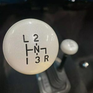 May include: White gear shift knob with black lettering indicating the gear positions: L, 2, 1, N, R, 3. The knob has a textured surface.
