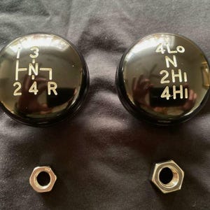 May include: Two black round gear shift knobs with white lettering. The knob on the left has the numbers 1, 2, 3, and 4, the letters 'R' and 'N', and an arrow pointing to the left. The knob on the right has the numbers 2 and 4, the letters 'N', 'H', and 'L', and the text '4LO', '2HI', and '4HI'.
