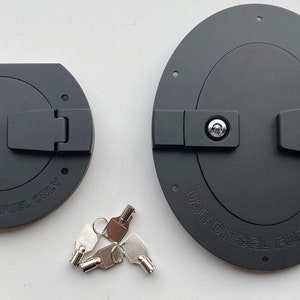 Puede incluir: Dos tapas de tanque de combustible de plástico negro con mecanismos de bloqueo y llaves. La tapa más grande tiene el texto "USAR SOLO COMBUSTIBLE DIESEL" y la tapa más pequeña tiene el texto "COMBUSTIBLE FUERA DE CARRETERA SOLO".