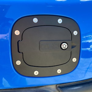 Puede incluir: Una tapa de gasolina negra con una cerradura plateada y la palabra "TRD" en relieve. La tapa está rodeada por un anillo de tornillos plateados y está montada en un coche azul.