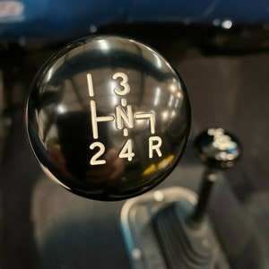May include: Close-up of a black gear shift knob with white numbers 1, 2, 3, 4, R, and N. The knob is round and has a shiny finish.