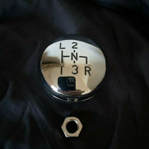 May include: A chrome gear shift knob with a black background and the letters L, 2, R, N, 1, and 3 engraved on the top.